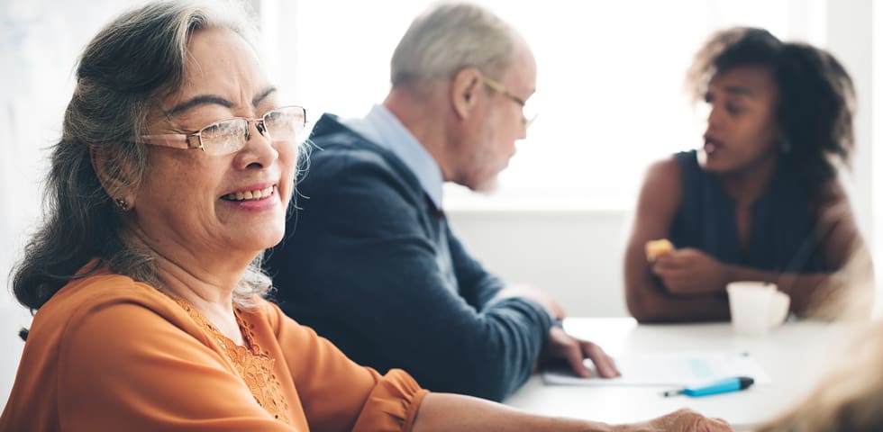 An older lady wearing an orange top and wearing glasses sits at a boardroom table. She is smiling and there are two people in the background.